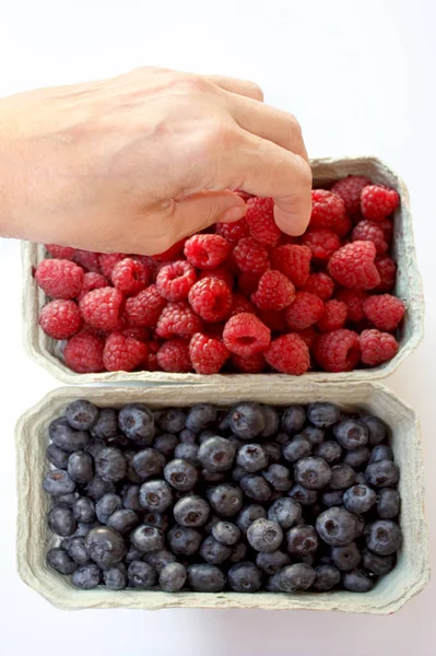 Raspberry and blueberry in packing containers, womans hand takes berries from box, red and blue fruit in package, closeup.