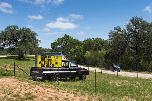 Pareja en una motocicleta pasando por un camión con un cartel para un evento musical en Luckenback, Texas . —  Fotos de Stock
