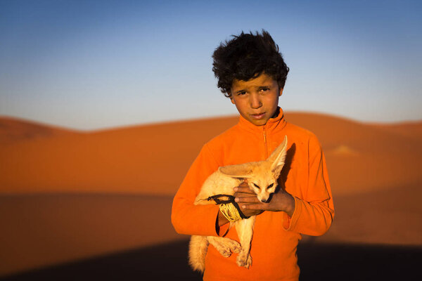 Berber child holding a desert fox poses in the Erg Chebbi dunes in Morocco.