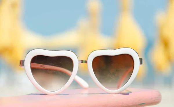 Μόδας γυαλιά ηλίου καρδιά σχήμα σε παραλία φόντο με κίτρινα ομπρέλες — Φωτογραφία Αρχείου