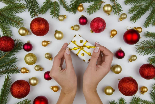 Composição de Natal feita de bolas de Natal douradas e vermelhas, cones e ramos de abeto. Em fundo branco mãos femininas segurando uma pequena caixa de presente. Inverno flat lay — Fotografia de Stock