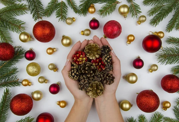 Composição de Natal feita de bolas douradas e vermelhas para uma árvore de Natal, cones e ramos de abeto. Em fundo branco mãos femininas segurando cones de Natal. Inverno flat lay — Fotografia de Stock