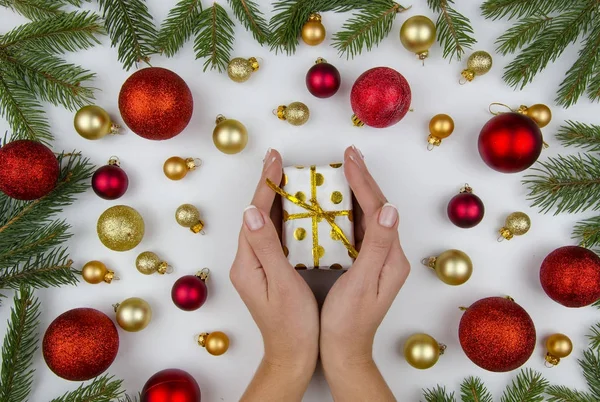 Mãos femininas segurando uma pequena caixa de presente no fundo branco com bolas de Natal douradas e vermelhas e ramos de abeto. Inverno deitado. Composição de Natal . — Fotografia de Stock
