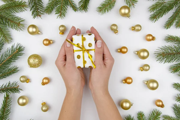 Christmas sammansättning av gyllene julgranskulor, kottar och fir grenar. På vit bakgrund kvinnliga händer som håller en liten presentbox. Vinter platt låg — Stockfoto