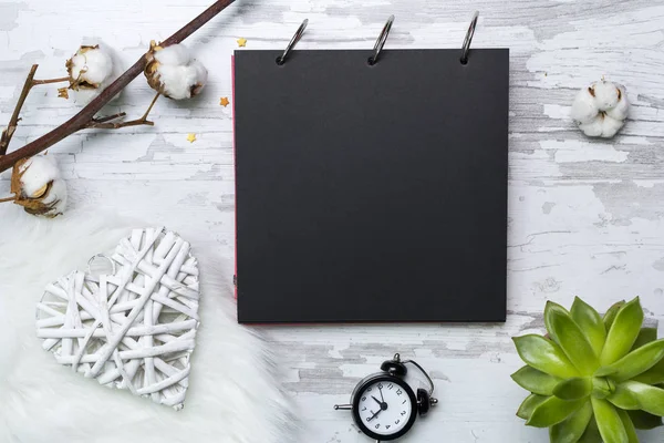 Black paper notebook, white heart, alarm clock,green plant and cotton flower. Flat lay mockup for inspiration quote about time, love and life.