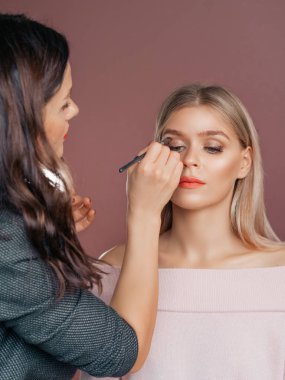 Dikey makyaj sanatçısı, bej arka planda makyaj yapan güzel sarışın bir kadına makyaj yapar.