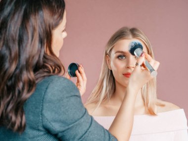 Profesyonel makyöz işyerinde güzel sarışın kadın makyajı yapıyor. Sahne arkası pembe renkler