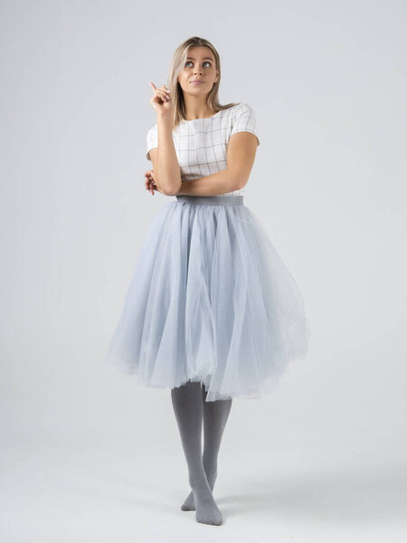 Vertical shot of amazed blonde girl model have an idea on cyclorama white background in a studio. Hand with finger pointing up. Full growth in profile in pastel colors. Long skirt and gray tights