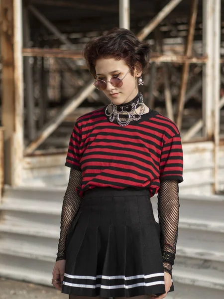 Punk woman cutted body on an abandoned construction background. A teen girl in a skirt, a collar and round glasses looks down thoughtfully