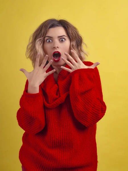 Surprise expression shock portrait of beautiful woman looking at camera in a red sweater on a yellow background. Demonstration of wow effect