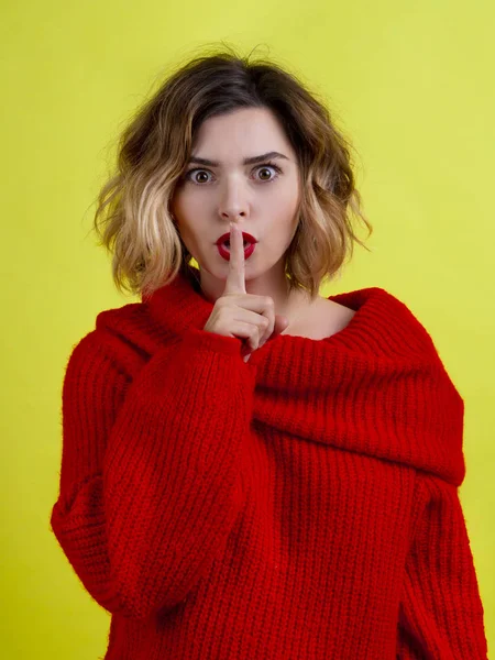 Sexy woman in red asking for silence and secrecy with a finger on her red lips. Hand gesture isolated on yellow background. Facial expression
