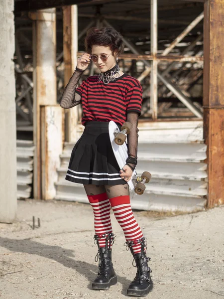 Menina alternativa em um fundo de construção abandonada decolando hipster óculos redondos e detém skate branco penny — Fotografia de Stock