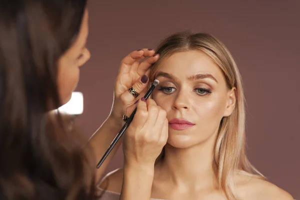 An artist applying eyeshadow powder with a brush for smokey eyes makeup on a pretty woman. Face close-up. Makeup lessons