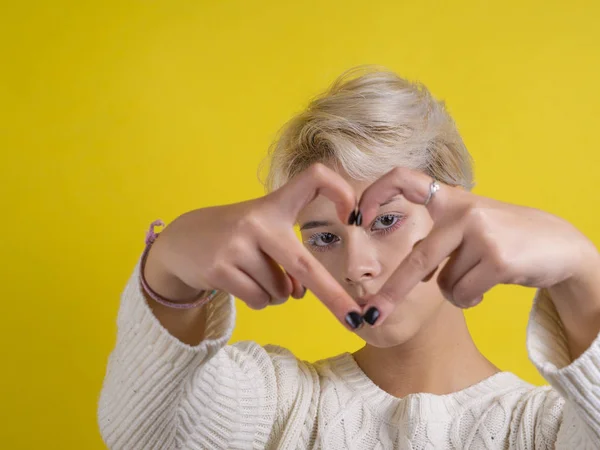 Retrato alternativo adolescente chica sobre fondo amarillo haciendo signo de corazón con los dedos y mira a través de ella . —  Fotos de Stock
