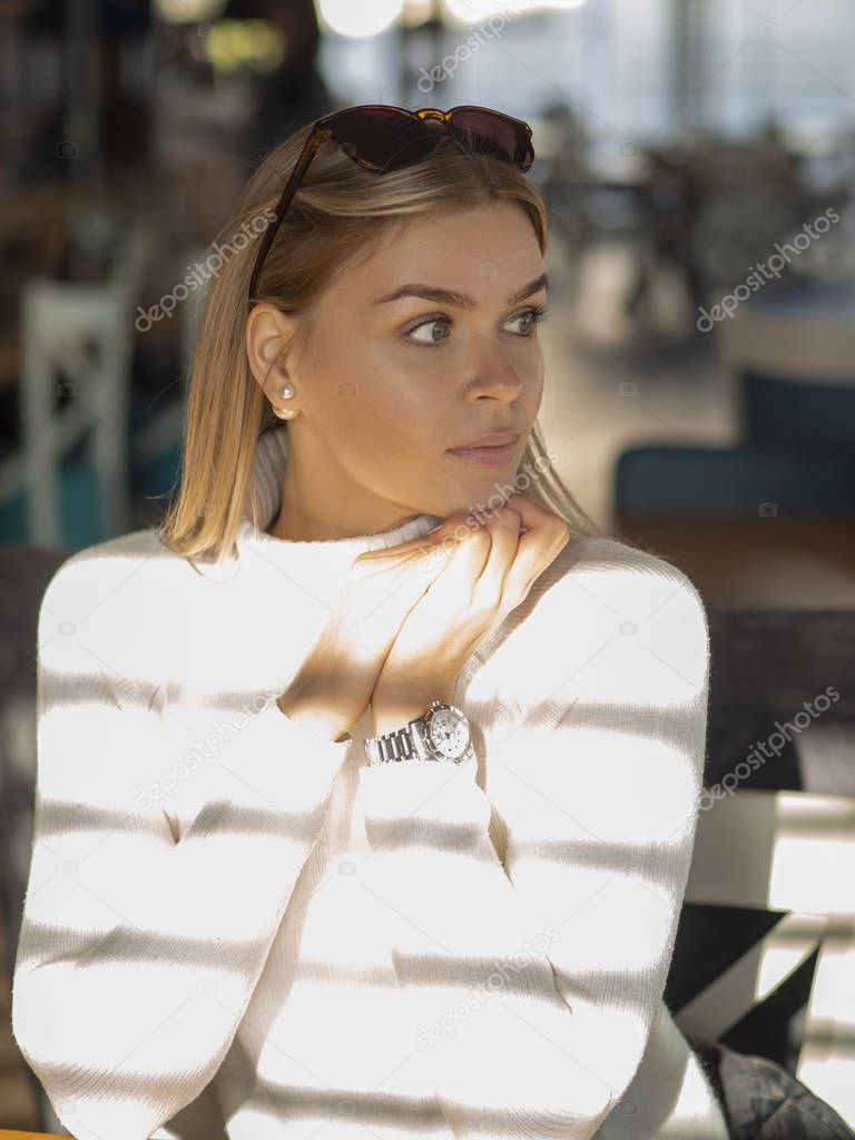 Beautiful woman looking away and sitting in a cozy cafe in a white sweater, the suns rays fall on her
