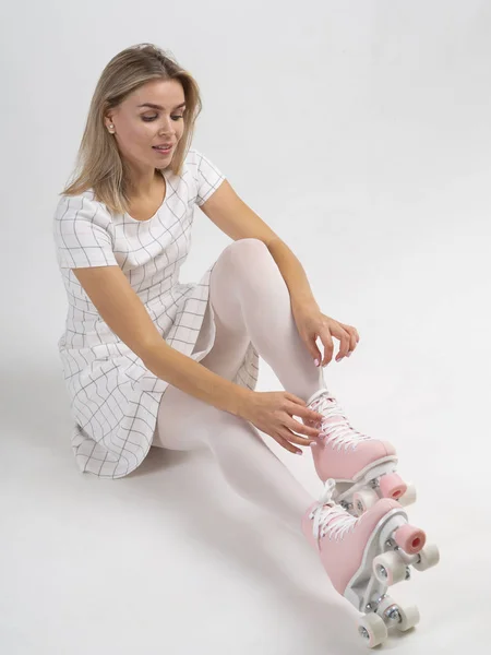 Prepare to skate. Cheerful young woman coquettishly tying shoelaces on her vintage quad wheels roller skates while sitting on the floor against a white background