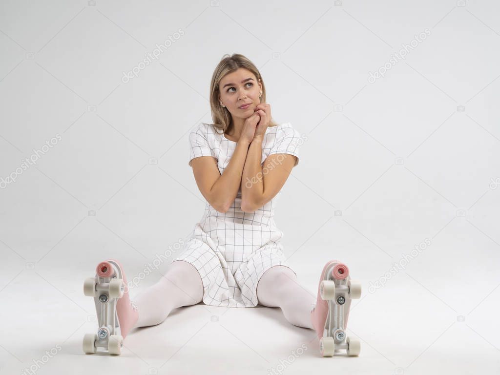 Joyful blonde woman in white with pink retro roller skates shoes sitting on the floor and dreaming. Looking up, hands touching necks. Idea for a banner. Dreaming concept