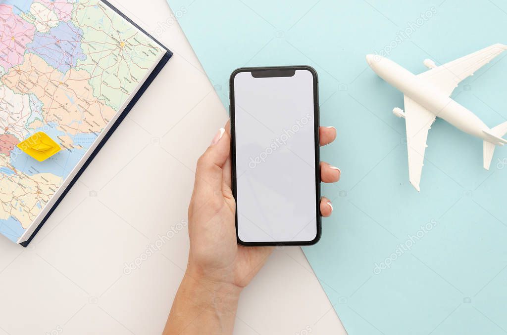 Top view mockup womans hand holding the black smartphone with an empty screen. Travel flat lay with map and airplane toy