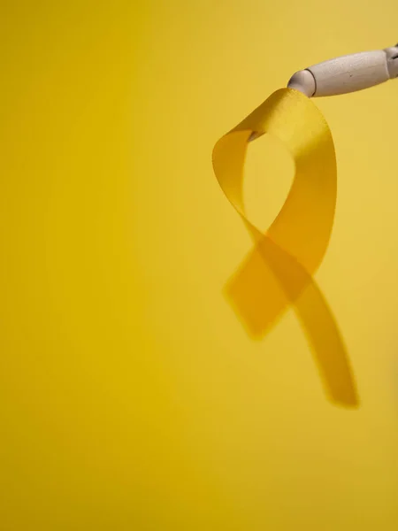 Wooden doll toy holds Childhood Cancer Awareness Gold Ribbon on yellow background. Close Up vertical still life photo — ストック写真