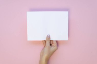Yukarıdan kadın eli görüntüsü, pastel pembe arka planda boş kağıt örtüsü tutuyor.