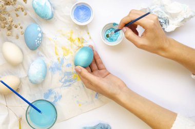 Easter eggs painting in blue colors by the woman on a white background. Feminine hands holds paintbrush and paints on an egg