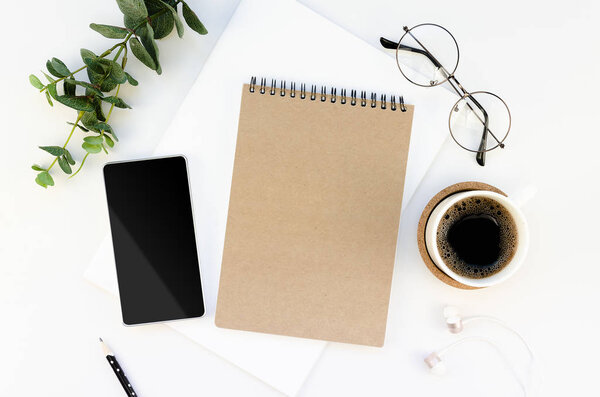 Feminine workspace mockup. Blank kraft paper spiral notepad and black screen smartphone and cup of coffee. Bordered with Eucalyptus and glasses