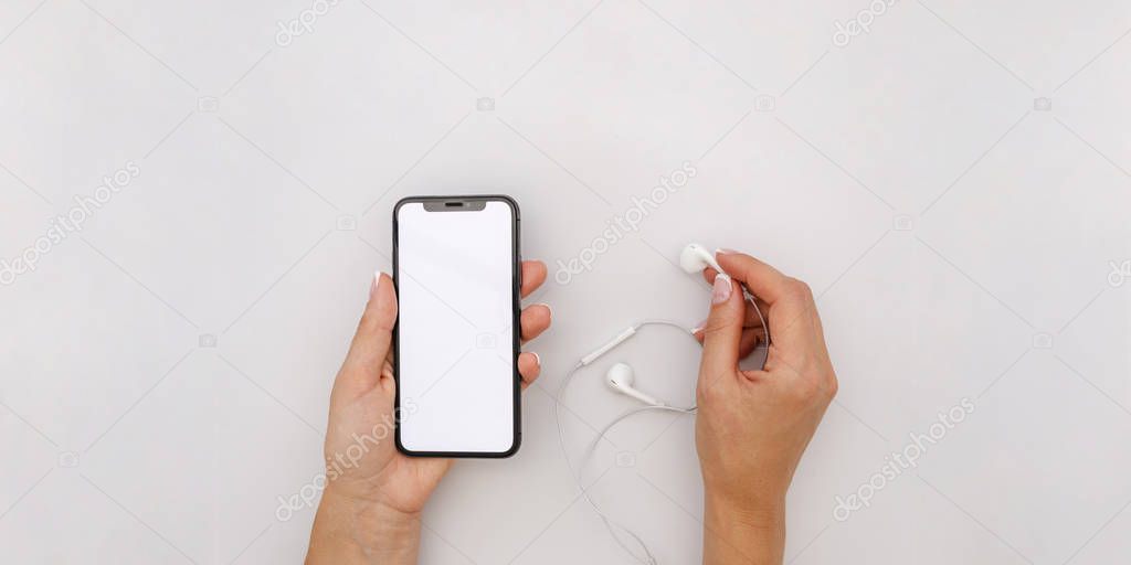 Feminine hands holding earphones and the black iphone x with white screen on a grey background. Top view