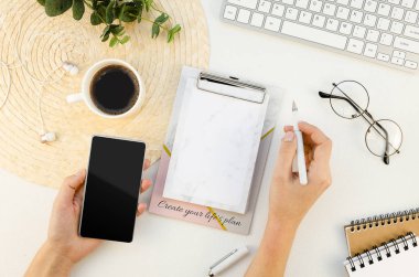 İş kadını beyaz bir masada klavyeyle çalışıyor, akıllı telefon model ekranını tutuyor ve not alıyor. Kopyalama alanı olan üst görünüm, düz yatay