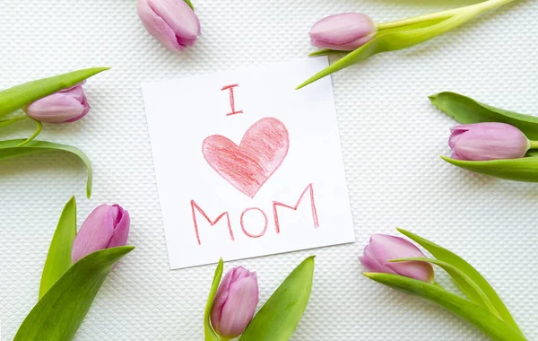 Plano lay childs dibujo amo a mi mamá. Enmarcado con tulipanes sobre una mesa blanca. Día de las madres y concepto familiar — Foto de Stock