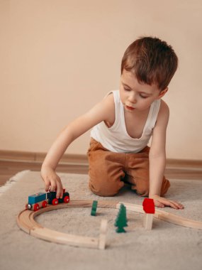 Küçük çocuk yerde oturup ahşap bir tren oyuncağıyla oynarken ağzıyla çuf çuf tren yapıyor.