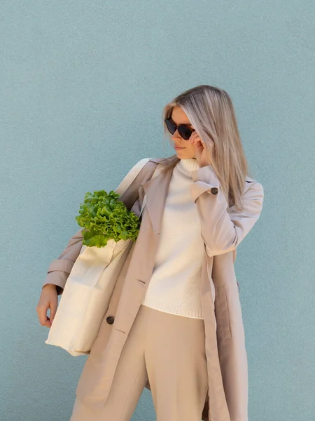 Elegante ama de casa que sostiene el bolso del supermercado del algodón con las verduras verdes frescas en un fondo azul de la pared. Toque gafas de sol y mirando hacia abajo — Foto de Stock
