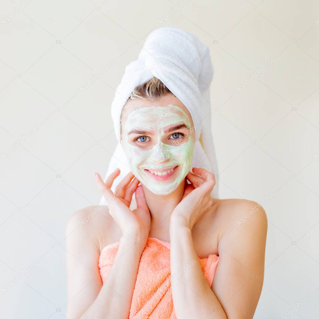 Beauty procedure. Portrait of a young woman with a towel on her head puts a nourishing cream on her face, she smiles and enjoys