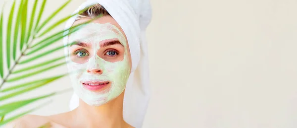 Wanita cantik dengan masker kecantikan hijau wajah dan handuk di kepalanya pada prosedur spa. Daun palem kabur. Spanduk lebar dengan salinan teks benteng spasi — Stok Foto