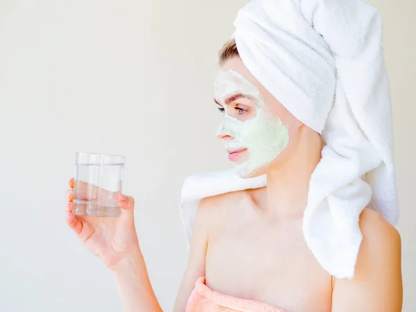 Agen pelembab alami H20 kosmetik bersih cocnept. Potret seorang gadis SPA dengan handuk di tubuh dan kepala dan masker wajah memegang air gelas bersih. Kulit wajah kering — Stok Foto