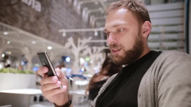 Homem bonito jovem com barba senta-se em um café, usa seu smartphone, sorri e acena. Tecnologia moderna . — Vídeo de Stock