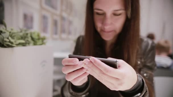 Mujer joven y bonita con el pelo oscuro en una chaqueta de cuero usando su teléfono inteligente, hablando, sonriendo. Fotos en la pared . — Vídeo de stock