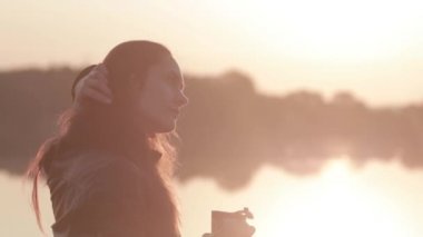 yakın çekim portre çekici genç kız güneş doğarken nehir ve içme kahve üzerinde bir thermocup görünüyor