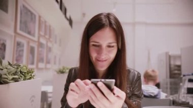 Onun smartphone kullanarak, söz, gülümseyen bir deri ceket siyah saçlı genç ve güzel kadın. Duvardaki resimler.