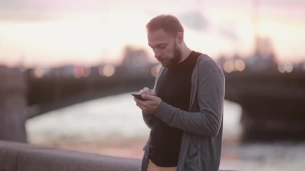 Homem bonito usando seu gadget. Homem usando um smartphone em pé em um cais. Carros ao fundo. Tecnologia moderna — Vídeo de Stock