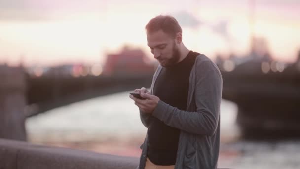 Un bel homme utilise un smartphone, regarde autour de lui. Un homme debout sur un quai. Voitures à l'arrière-plan. Technologie moderne — Video