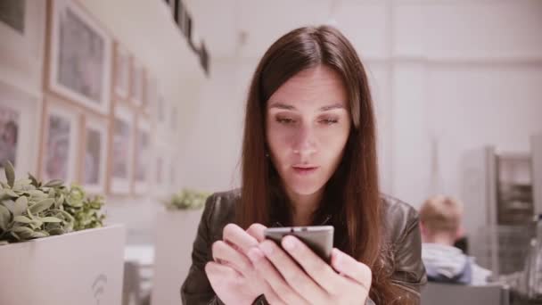 Bella giovane donna con i capelli scuri e gli occhi in una giacca di pelle utilizzando il suo smartphone. Stanza della luce, immagini sulla parete . — Video Stock