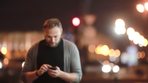 Stilig man promenader, ler, använder sin smartphone och går bort. Suddig stadens ljus på gatan. Modern teknik. — Stockvideo
