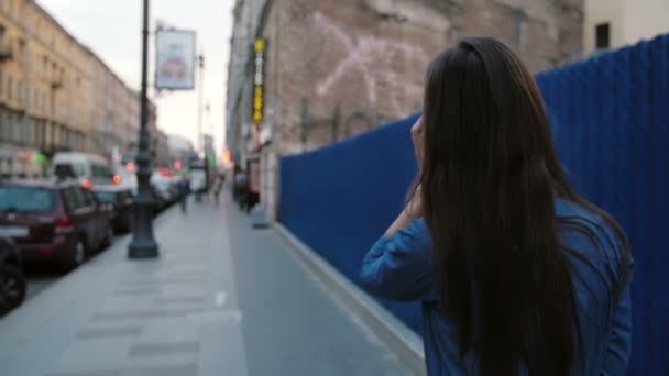 Frau läuft auf der Straße neben blauem Zaun. Rückansicht einer Frau mit langen Haaren, die am Telefon spricht. slow mo, steadicam shot — Stockvideo