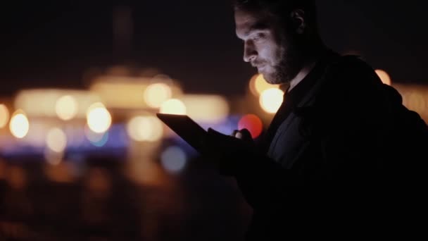 Bonito homem, sorri, acena, usa o tablet à noite, depois vai embora. Luzes turvas da cidade. Vista lateral. Tecnologia moderna — Vídeo de Stock