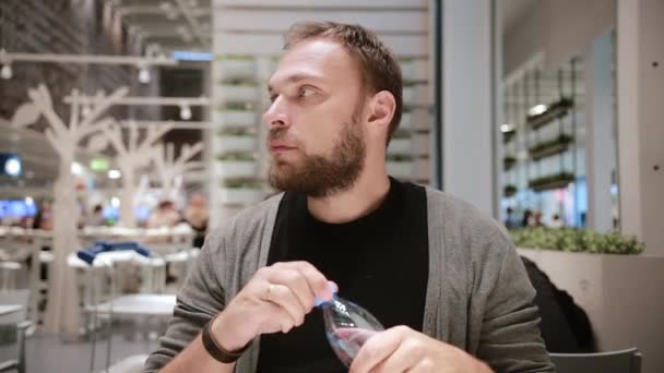 Trinkwasser. gutaussehender Mann mit Bart, der eine Wasserflasche öffnet, sitzt auf einem öffentlichen Platz. Mann hat Durst. — Stockvideo