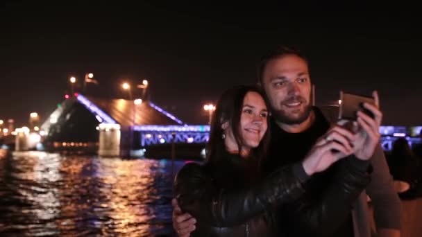 Um casal feliz apaixonado a tirar selfie. Uma ponte móvel do palácio São Petersburgo no fundo. Iluminação da cidade à noite — Vídeo de Stock