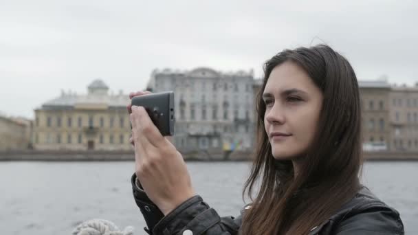 Una chica bonita toma fotos en una ciudad. Ella está contenta, sonriendo, usando su teléfono inteligente, arquitectura en el fondo, lento mo — Vídeo de stock