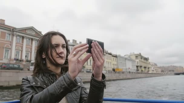 Jovem em um passeio pelo rio, tirando fotos, usando smartphone. O vento sopra o cabelo. Vista da cidade de São Petersburgo. Devagar. — Vídeo de Stock
