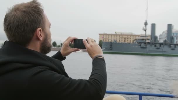 Um homem está a passear. Tirando fotos do cruzador Aurora em São Petersburgo, usando seu smartphone, backview, slow mo — Vídeo de Stock