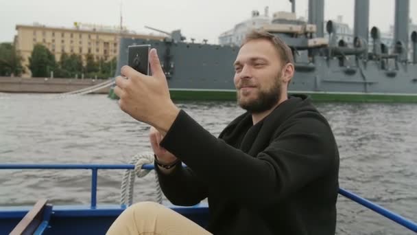 Счастливый красавец в Санкт-Петербурге. Делает селфи перед крейсером "Аврора", использует свой смартфон, медленное время — стоковое видео